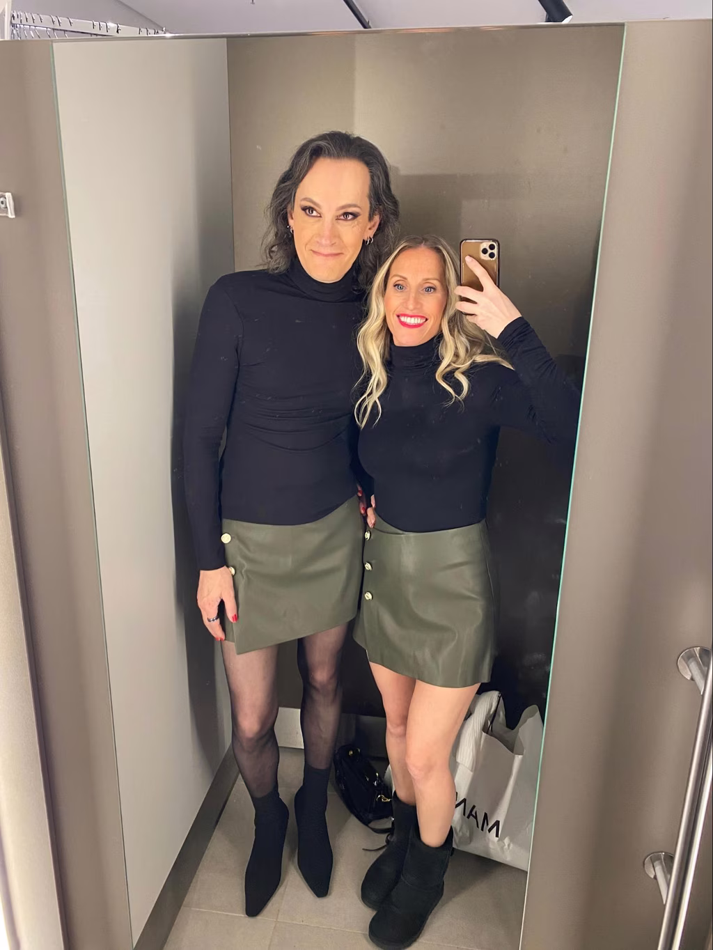 Le Duo en Jupe Verte Anaêlle et Jennifer apparaissent dans un selfie pris dans un miroir de cabine d'essayage. La personne à gauche porte un col roulé noir, une jupe courte de couleur olive avec des boutons dorés décoratifs sur le devant, des collants noirs semi-transparents, et des bottines noires. La personne à droite, tenant le téléphone, est habillée de manière similaire mais porte des bottes différentes.