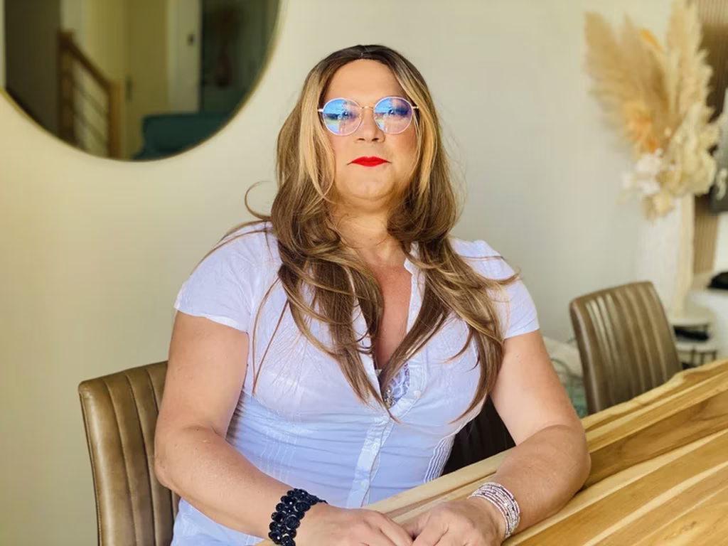 Laura, en séance de féminisation chez Transbeauté, explore avec grâce sa féminité. Assise à une table, elle porte une chemise blanche ajustée et des lunettes bleues, incarnant la dualité de son identité avec élégance et assurance