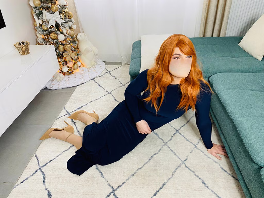 Travesti chez Transbeauté avec des cheveux roux portant une robe bleu foncé et des chaussures à talons hauts de couleur beige. Elle est assise sur un tapis, adossée à un canapé bleu-vert, avec un genou plié et l'autre jambe étendue. À côté d'elle, il y a un sapin de Noël richement décoré avec des guirlandes lumineuses, des ornements dorés et blancs, et une figurine d'ours polaire. La pièce semble lumineuse avec des murs blancs et des rideaux blancs transparents.