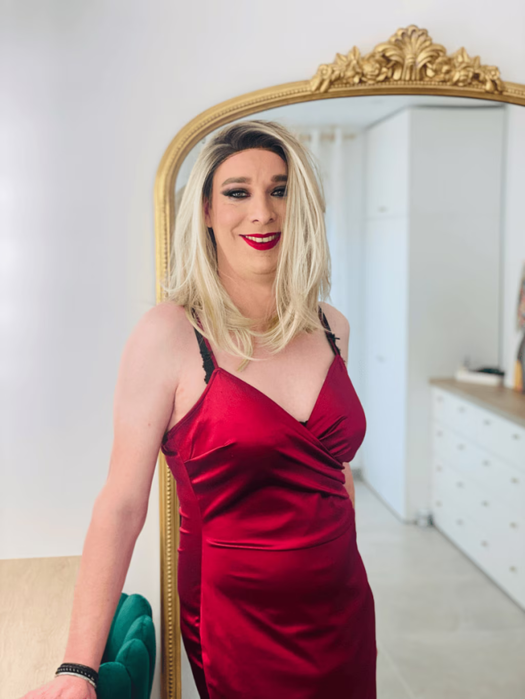 Stella en robe de satin rouge, coiffée et maquillée, dans un salon chic avec un grand miroir doré en arrière-plan