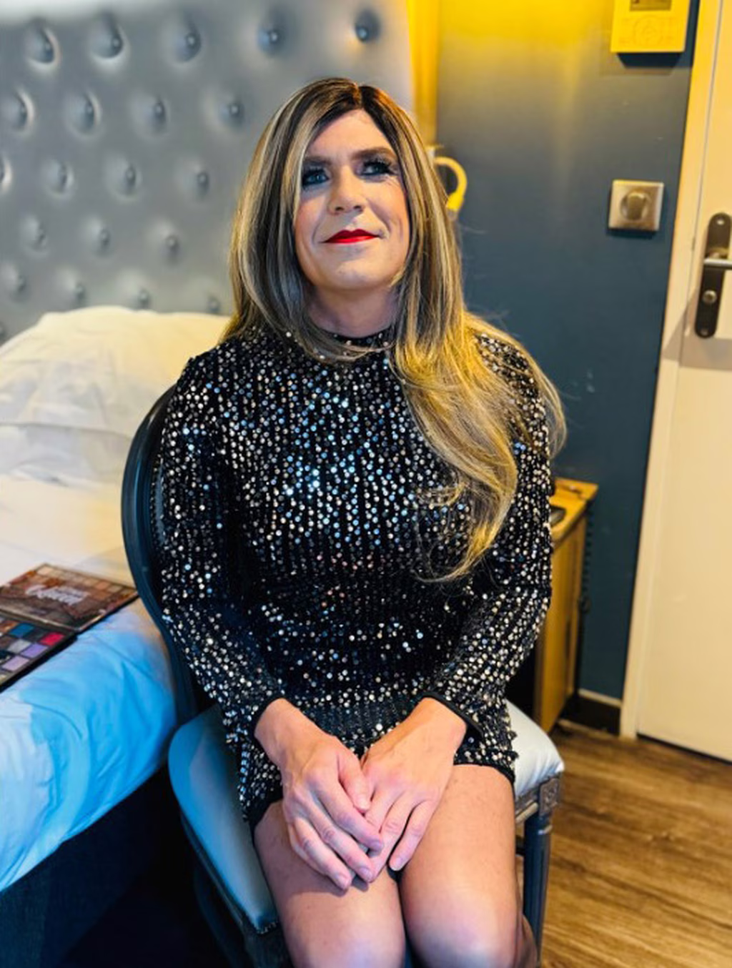 Stéphanie, habillée d'une robe noire à sequins, est assise dans une chambre d'hôtel, avec un sourire serein et les mains posées sur les genoux