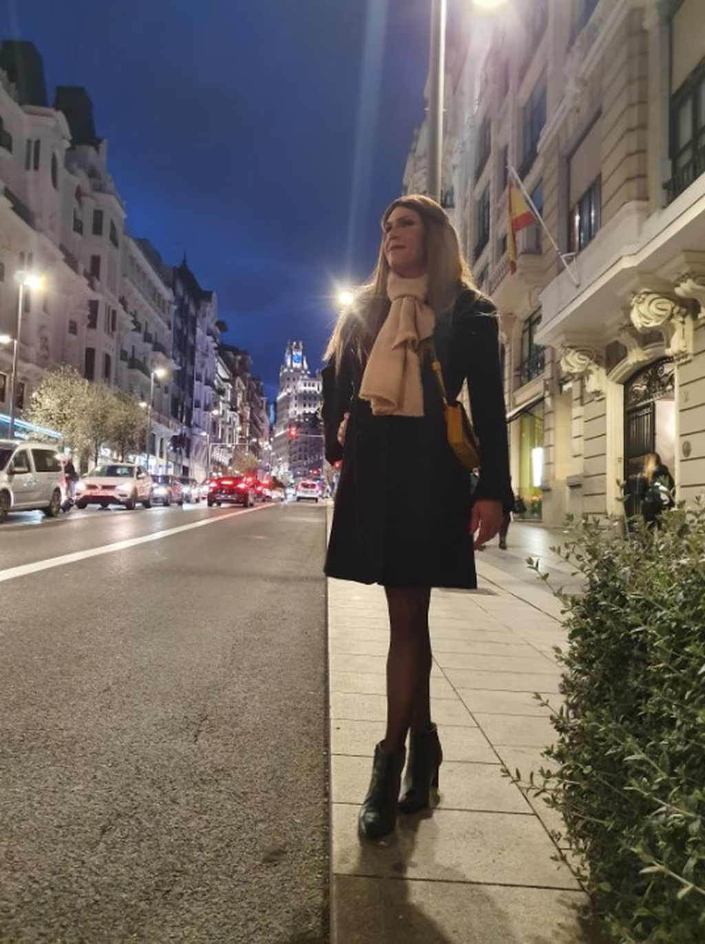 Stéphanie marche en soirée dans une rue animée, vêtue d'un manteau noir et d'une écharpe beige, avec des bottines noires. Elle est éclairée par les lumières urbaines, projetant une image de confiance et d'élégance dans cette scène nocturne