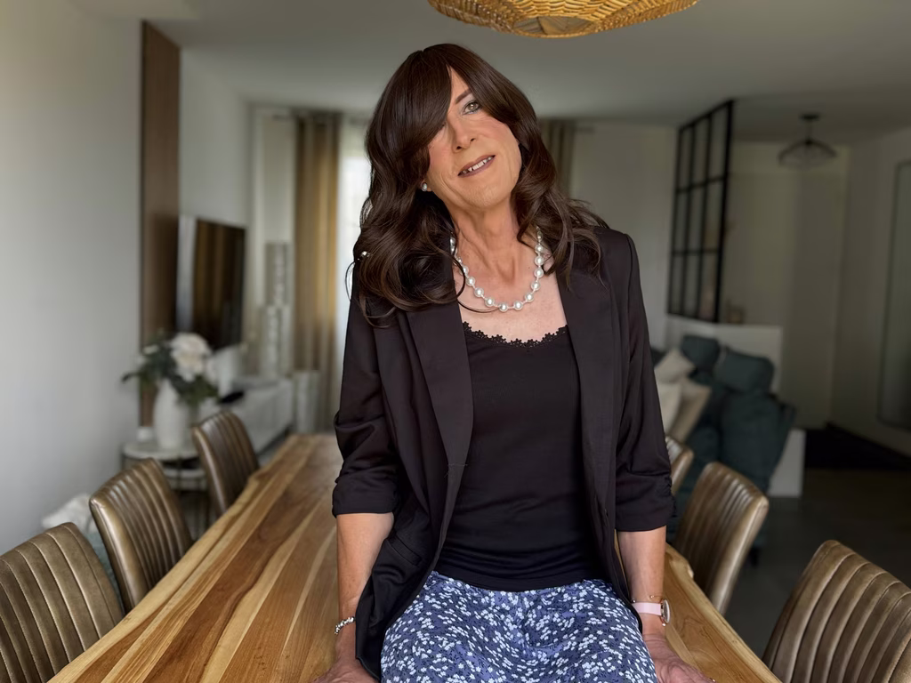 Femme assise sur une table en bois, souriante et confiante, vêtue d’un blazer noir élégant et d’un haut à dentelle, sublimée par un collier de perles et une coiffure soignée