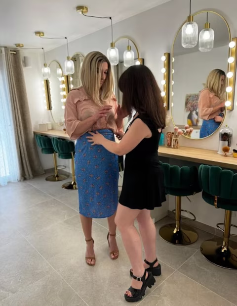 Stella, accompagnée de sa femme, vit un moment précieux chez Transbeauté. En suivant les conseils de Jennifer, la maquilleuse professionnelle, Stella se féminise avec élégance et confiance. Sa compagne l'accompagne et l'aide à ajuster sa tenue, renforçant leur lien de soutien et d'amour. Le salon, avec ses miroirs lumineux et son décor raffiné, reflète la beauté de ce moment de transformation. Stella, habillée d'une jupe bleue fleurie et d'une chemise rose, incarne pleinement son identité féminine, encouragée par l'approbation et le soutien inconditionnel de sa partenaire