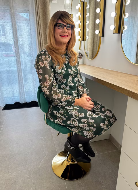 Amélie est assise devant une coiffeuse illuminée, portant une robe à motifs fleuris et des bottines noires élégantes. Son sourire et sa posture dégagent sérénité et assurance.