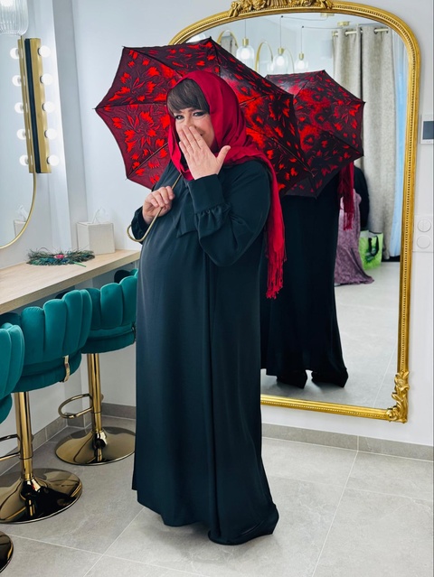 Femme trans musulman en robe noire et foulard rouge, posant avec espièglerie sous un parapluie rouge à motifs dans un décor élégant.