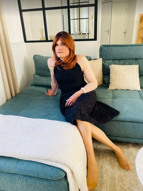 Carla, une personne transgenre, assise sur un canapé dans une robe noire, incarnant sa féminisation et son parcours de travesti avec élégance