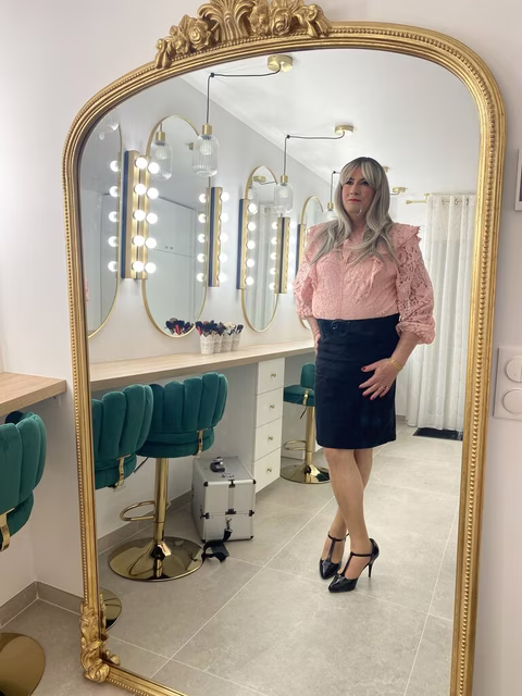 Chantal se tient avec élégance devant un majestueux miroir doré, son reflet ajoutant une dimension de beauté et de grâce. Vêtue d'un chemisier rose en dentelle et d'une jupe noire, ses cheveux gris argentés tombent en vagues délicates sur ses épaules. L'ambiance luxueuse de la pièce, avec ses miroirs éclairés et ses chaises en velours vert, amplifie l'aura de sophistication et de confiance qu'elle dégage. Ses mains posées sur ses hanches, elle incarne l'assurance et le charme intemporel