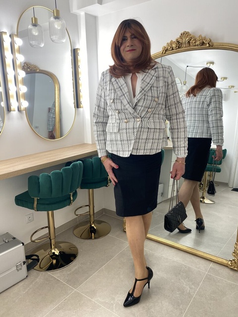 Chantal, habillée élégamment en veste en tweed à carreaux blancs et noirs et jupe noire, se tient debout dans une salle de maquillage chic. Elle porte des talons hauts noirs et tient un sac à main noir avec une chaîne argentée. Ses cheveux sont coiffés en une coupe mi-longue rousse. La salle est décorée avec des chaises vertes en velours et des miroirs dorés, créant une ambiance luxueuse