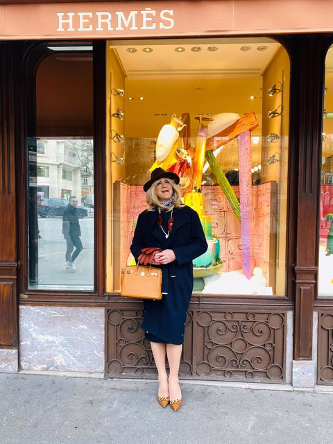 Deborah posant avec élégance devant une vitrine Hermès, vêtue d’un manteau bleu marine, d’un chapeau chic et d’accessoires sophistiqués.