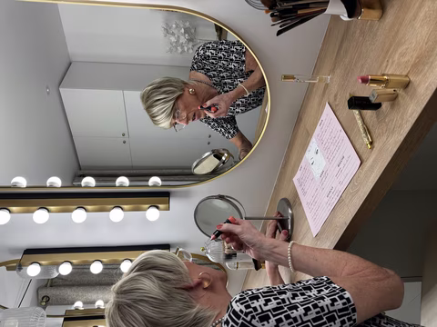 Une personne transgenre ou travestie applique soigneusement du maquillage devant un miroir illuminé. L'espace est aménagé avec des produits de beauté et des outils de maquillage, reflétant une routine soignée et affirmée