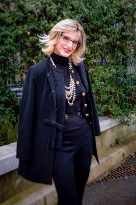 Glawdys souriante dans une tenue noire élégante, composée d'un manteau structuré, d'un pull col roulé et de bijoux dorés pour une touche sophistiquée