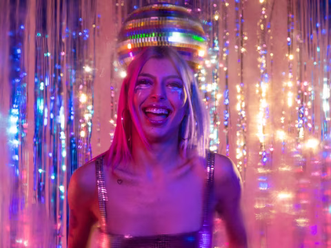 ne personne joyeuse et souriante dans une ambiance de fête, avec une boule à facettes suspendue au-dessus, reflétant les lumières multicolores qui l'entourent. Elle porte une tenue scintillante, et l'arrière-plan est orné de guirlandes lumineuses et de rideaux métalliques, créant une atmosphère éclatante et festive