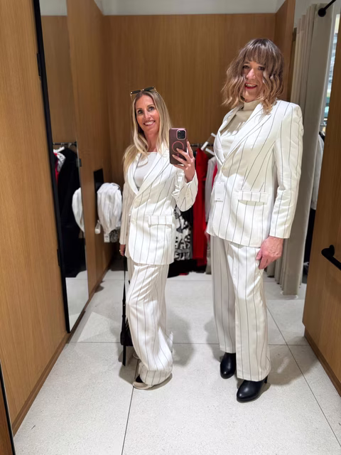 Jennifer, maquilleuse de Transbeauté, pose avec une cliente dans une cabine d’essayage. Toutes deux portent un tailleur blanc élégant, symbole de féminité et de confiance.