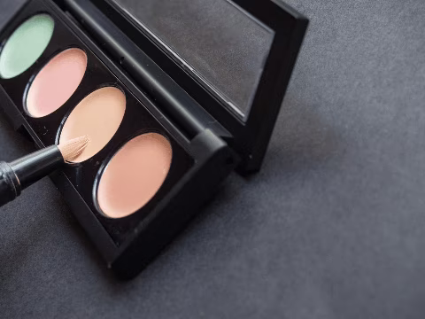 Une palette de correcteurs de couleurs avec quatre teintes, comprenant du vert pour neutraliser les rougeurs, du rose pâle pour illuminer, et deux nuances de beige pour unifier le teint. Un petit pinceau est utilisé pour prélever le produit. Cette palette est idéale pour les personnes transgenres ou travesties qui cherchent à créer une base uniforme et naturelle avant d'appliquer d'autres produits de maquillage