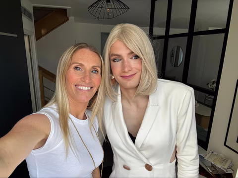Jennifer et Mélissa prennent un selfie dans un intérieur lumineux. Mélissa, grande, rayonnante dans une robe blazer blanche, affiche un sourire timide mais sincère. Elle se tient proche de Jennifer, qui sourit à l’objectif, confiante et complice. On sent la fierté douce, le moment suspendu avant de franchir une nouvelle étape.