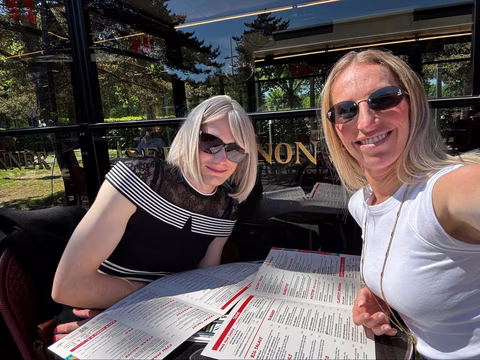 Mélissa et Jennifer sont attablées en terrasse, lunettes de soleil sur le nez, menus en main. Mélissa porte une robe noire à rayures et dentelle, très élégante, et semble parfaitement à l’aise. Jennifer, fidèle à elle-même, affiche son sourire franc. L’ambiance est détendue, complice, et joyeusement « normale » — ce genre de moment simple qui veut dire beaucoup.
