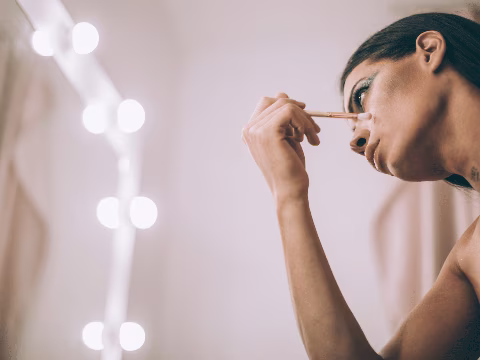 Une personne en train de se maquiller devant un miroir éclairé par des ampoules. Elle tient un pinceau et applique du maquillage sur son visage, se concentrant sur ses yeux. L'ambiance de la pièce est chaleureuse, avec une lumière douce qui ajoute à l'atmosphère intime de la scène. Le reflet des lumières crée une esthétique de salle de maquillage professionnelle