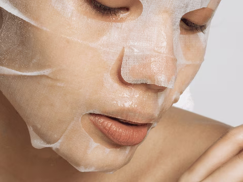 Une personne porte un masque facial en tissu, indiquant un moment de soins personnels et de détente. Le masque imprégné d'un produit hydratant illumine la peau, suggérant une étape de préparation essentielle avant le maquillage. Cette image met en valeur l'importance des soins de la peau pour obtenir un teint frais et éclatant, une pratique cruciale pour les personnes transgenres ou travesties qui souhaitent un maquillage impeccable