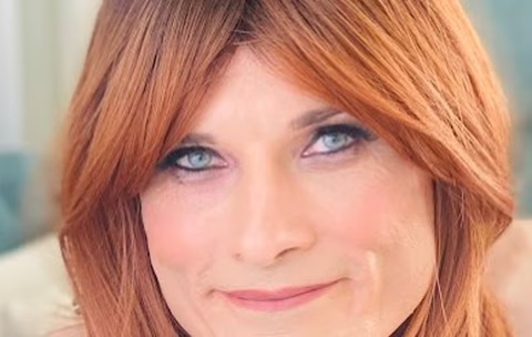 Portrait rapproché d'une femme transgenre aux cheveux roux, maquillée avec élégance, regardant doucement la caméra avec un sourire apaisant