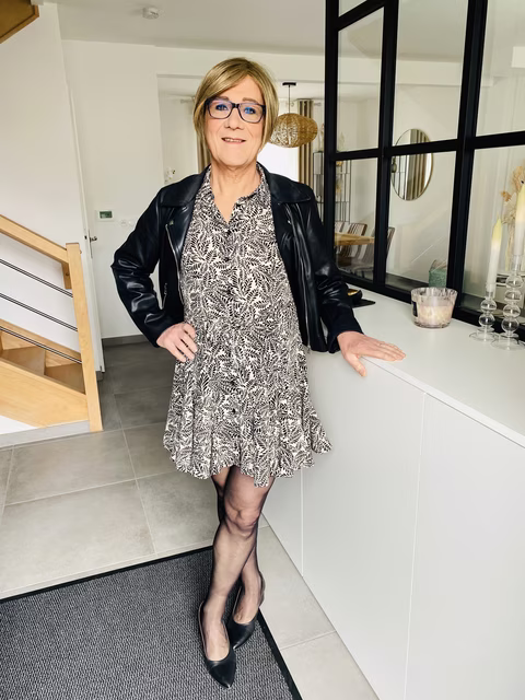 Roxanne, souriante et vêtue d'une robe à motifs noirs et blancs avec une veste en cuir noire, pose dans une pièce moderne et lumineuse