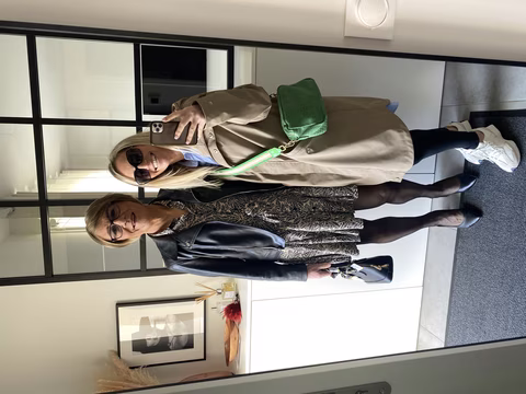 Roxanne, vêtue d'une robe à motifs et d'une veste en cuir, pose avec Jennifer, qui porte un trench-coat beige et des baskets blanches, pour un selfie dans une pièce lumineuse