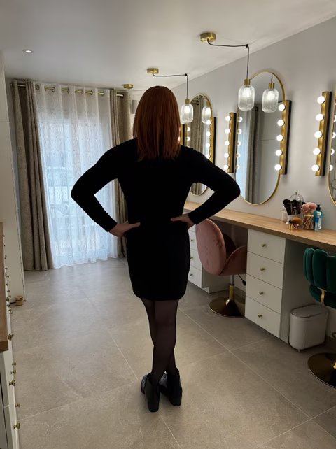 Une personne élégante en robe noire, vue de dos, pose dans un studio beauté lumineux et raffiné. Les miroirs éclairés et les fauteuils en velours créent une atmosphère professionnelle et accueillante. Cela pourrait bien correspondre à une séance réalisée chez Jennifer de Transbeauté, où l'accent est mis sur l'exploration de l'identité féminine et le soin de soi, comme dans le cas de Roxanne, travesti ou transgenre en pleine expression de sa féminité