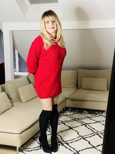 Sabine, debout et souriante, dans un salon lumineux. Elle porte une robe-pull rouge vif, qui contraste joliment avec sa peau et lui arrive mi-cuisse, mettant en valeur ses jambes complétées par des collants noirs et des chaussures à plateforme de la même couleur. Sa tenue dégage une sensation chaleureuse et confortable tout en ajoutant une touche de couleur éclatante. Ses cheveux blonds sont coiffés en une frange droite et tombent librement un peu en dessous de ses épaules. Son maquillage est discret mais rehausse ses traits, avec une attention particulière portée à ses yeux et ses lèvres. Le décor derrière Sabine est neutre et moderne, avec un canapé beige en forme de L et un tapis à motifs géométriques. La pièce semble être bien éclairée, avec une lumière naturelle qui entre par les fenêtres, renforçant l'atmosphère accueillante de l'espace de vie.