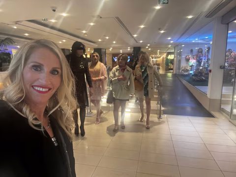 Selfie de Jennifer dans le hall d’un hôtel avant la soirée Transbeauté, avec plusieurs participantes travesties ou transgenres en tenues élégantes, maquillages soignés et looks festifs
