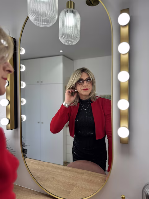 Résultat d’une séance maquillage Transbeauté : femme travestie élégante devant un miroir éclairé, robe noire et veste rouge, maquillage sublimé et regard confiant.