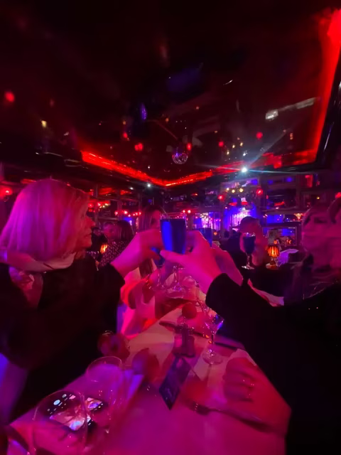 L’ambiance est festive et chaleureuse. On y voit un petit groupe de femmes attablées dans un restaurant ou un cabaret au décor tamisé, baigné de lumières rouges et violettes. Les verres se lèvent pour un toast : un moment de partage, de complicité et de célébration. Les regards sont bienveillants, les sourires discrets mais sincères. L’atmosphère, entre glamour et convivialité, évoque une soirée où chacune peut être pleinement elle-même — élégante, libre et rayonnante. Un instant capturé qui résume bien l’esprit Transbeauté : féminité, confiance, et joie d’être ensemble