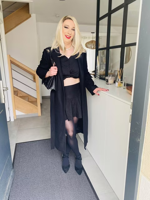 Stella, une personne travestie, se tient debout dans un intérieur moderne, souriante, portant une tenue chic tout en noir. Elle arbore un crop top, une jupe, des collants noirs, et des bottines pointues. Un manteau long et une pochette en chaîne ajoute une touche élégante à son look. Elle pose dans un couloir lumineux, avec une décoration intérieure épurée et moderne en arrière-plan