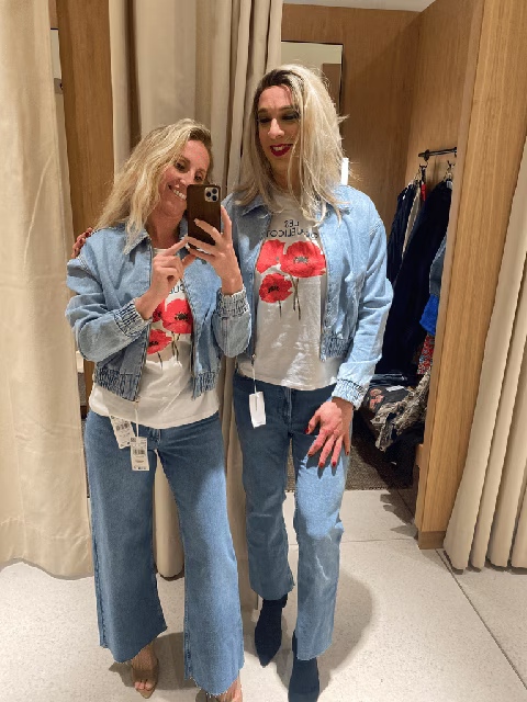 Image de Jennifer, maquilleuse professionnelle spécialisée pour les personnes travesties et/ou transgenres, lors d'une sortie shopping. Jennifer accompagne une de ses clientes, Stella, pour une séance d'essayage en magasin. Elles portent des tenues assorties et posent joyeusement pour un selfie dans la cabine d'essayage, illustrant le soutien et la confiance que Jennifer apporte à ses clientes lors de ces expériences sociales enrichissantes
