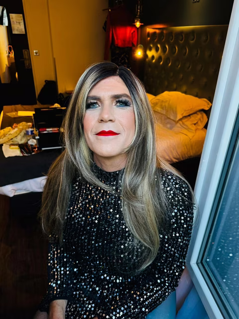 Stéphanie, en robe noire à sequins, est assise près d'une fenêtre, affichant un maquillage élégant et un regard confiant dans une chambre d'hôtel au décor chaleureux
