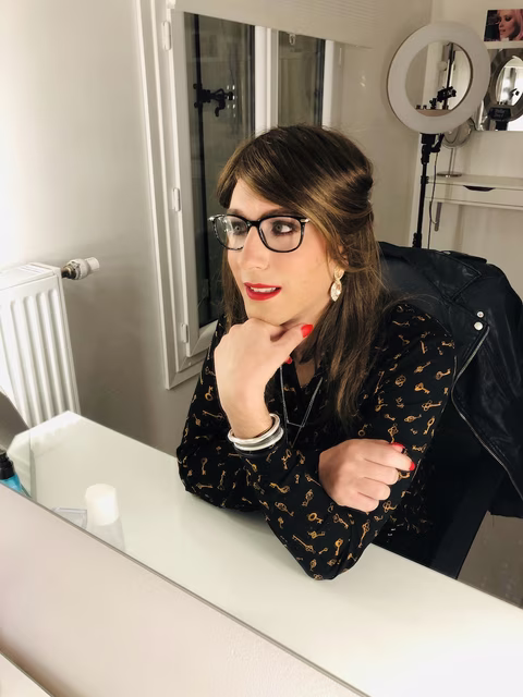 Femme transgenre assise devant un miroir, portant des lunettes et une blouse noire à motifs dorés. Elle adopte une pose confiante, la main sous le menton, et semble se contempler attentivement