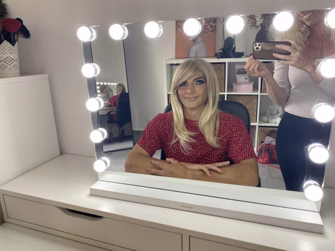 Vanessa femme travestie assise joyeusement devant un miroir de maquillage, portant une perruque blonde stylée et un haut à pois. Une atmosphère d'échange et de métamorphose semble régner, avec Jennifer en arrière-plan participant au processus de transformation, créant une ambiance d'accompagnement et de créativité